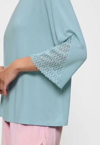 Jasnoniebieska bluzka z rękawami o długości trzech kwart, ozdobiona koronkowym wykończeniem przy mankietach. Miękka tkanina i luźny krój, noszona na różowych spodniach.