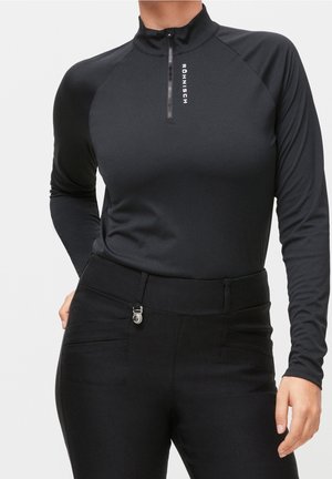 Femme portant un haut noir ajusté à manches longues avec une fermeture éclair au quart et un logo, associé à un pantalon noir taille haute avec une boucle métallique.