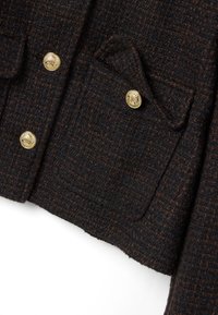 Donkerbruine getextureerde tweedjas met goudkleurige metalen knopen met reliëf en een klepzak aan de onderkant aan de voorkant.