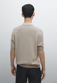 Polo a maglia beige con maniche corte e colletto a costine, caratterizzato da una superficie testurizzata. Indossato con pantaloni scuri, visto da dietro.