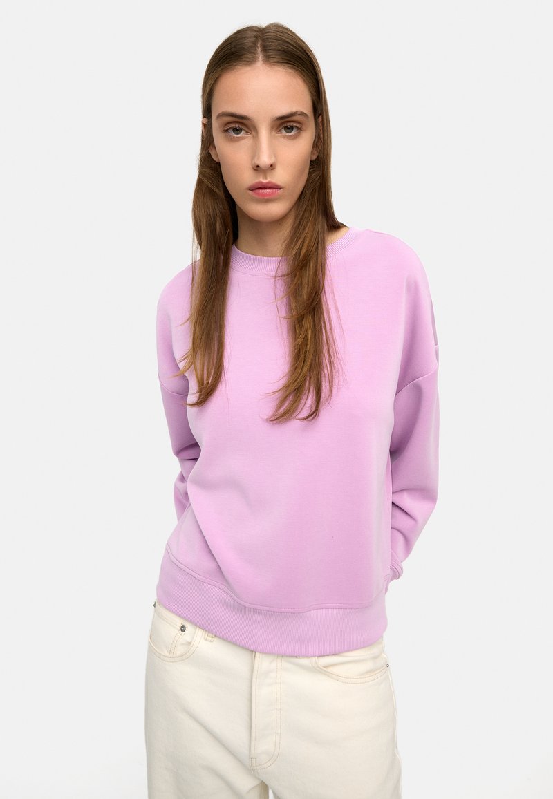 Młoda kobieta o długich brązowych włosach, ubrana w jasnofioletową bluzę i kremowe spodnie, stoi na tle gładkiego jasnego tła.