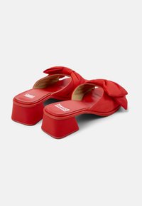 Red platform mules with a bow detail, made of smooth material. Square heel and open toe, featuring a cushioned insole and branding on the footbed.
