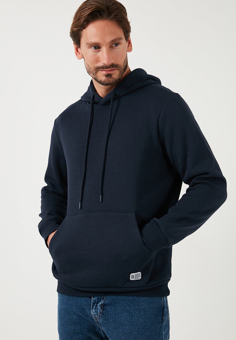 Felpa blu navy realizzata in tessuto morbido, con cappuccio regolabile, tasca frontale e una piccola etichetta con il marchio in basso a sinistra.