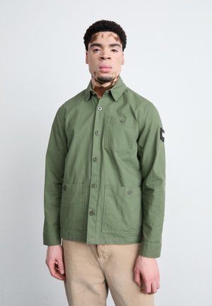 Khaki green jacket with a button front, two front pockets, and a patch on the left sleeve. Cotton material, straight cut, and long sleeves.