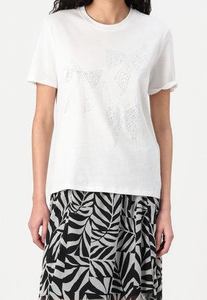 Biała bawełniana koszulka z trzema błyszczącymi kokardkami ozdobionymi cyrkoniami. Dół prosty, z podwiniętymi rękawami. W połączeniu z czarno-szarą spódnicą w liściasty wzór.