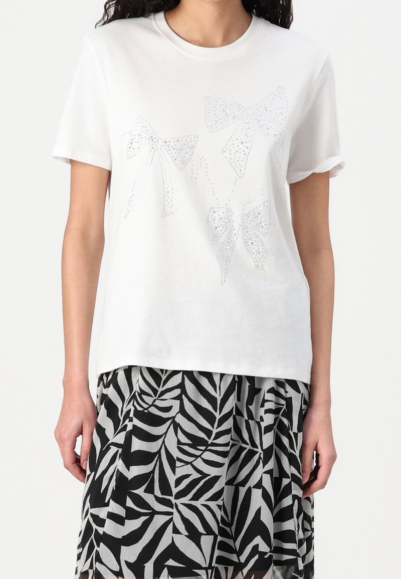 T-shirt en coton blanc avec trois motifs de nœuds brillants ornés de strass. Ourlet droit avec manches retroussées. Associé à une jupe imprimée de feuilles noires et grises.