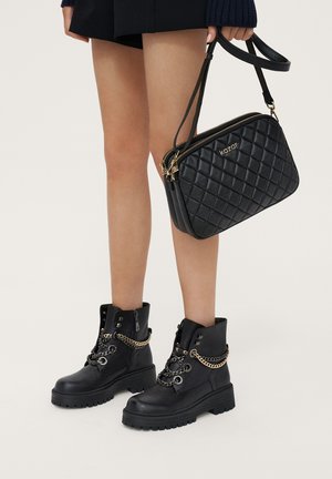 Bottines noires avec des accents en chaîne, en cuir texturé et à semelles chunky, assorties à un sac à bandoulière noir matelassé avec une fermeture éclair dorée.