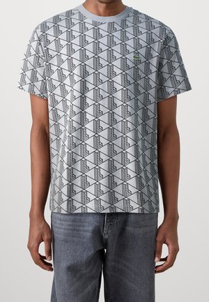 T-shirt en coton gris avec un motif géométrique noir. Col rond avec manches courtes. Logo Lacoste vert sur la poitrine gauche.