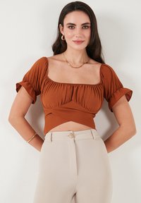 Rustfarvet cropped top med korte puffærmer, smocked detalje omkring udskæringen og en asymmetrisk foran kant. Båret sammen med beige bukser.
