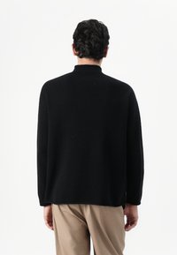 Maglione nero a maniche lunghe con collo alto, realizzato in un tessuto morbido. Presenta una vestibilità rilassata e un orlo dritto, indossato sopra pantaloni beige.