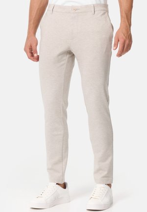 Man die licht beige slim-fit broek en witte lage sneakers draagt, staand tegen een effen achtergrond.