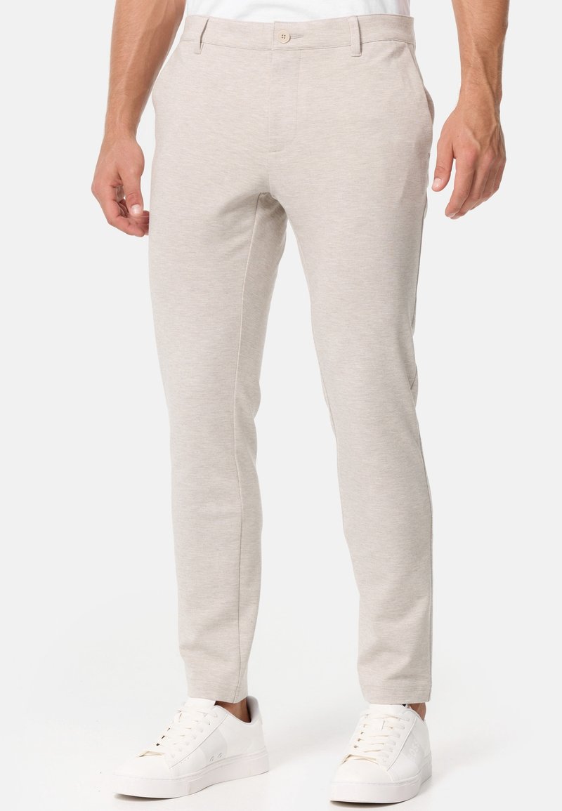 Homme portant un pantalon slim beige clair et des baskets basses blanches, debout devant un fond uni.