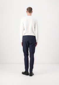 White, textured long-sleeve top paired with navy tailored trousers. Model shown from the back, wearing black shoes with a smooth finish.
