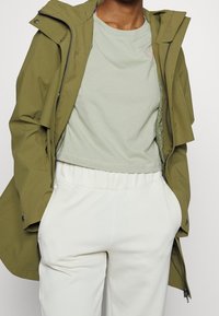 Oliwkowa kurtka przeciwwiatrowa z zamkiem z przodu, nałożona na jasną zieloną skróconą koszulkę, zestawiona z białymi spodniami na gumce.