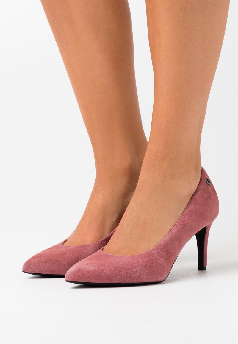 Jambes portant des escarpins roses en daim à bout pointu avec des talons fins, photographiés sur un fond blanc uni.