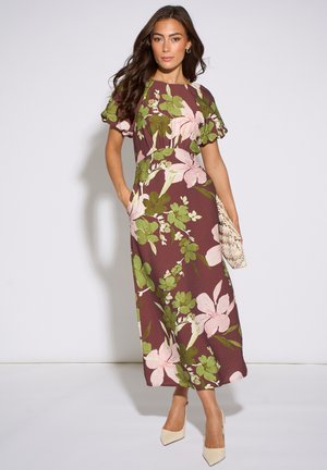 Woman wearing brown midi dress with pink and green floral print, holding a woven clutch and wearing beige pointed heels.