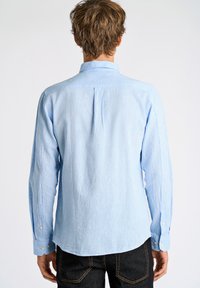 Camisa de manga larga azul claro hecha de un tejido texturizado. Presenta un cuello abotonado y un pliegue en la parte posterior. Combinada con jeans oscuros.