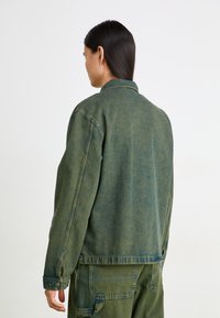 Jachetă din denim verde cu guler, cusături vizibile și buzunare laterale. Prezintă o textură ușor decolorată și o croială relaxată.
