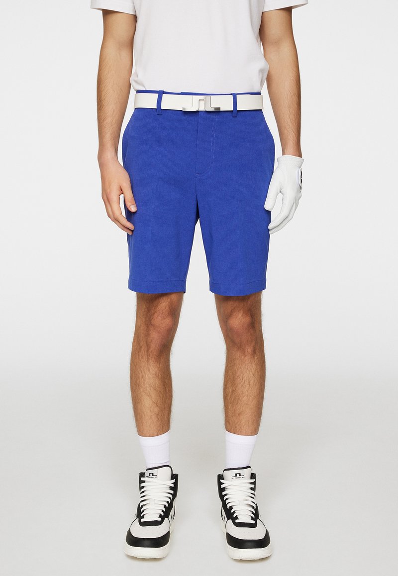 Blå golfshorts lavet af struktureret stof, med et hvidt bælte og en forlommekasse, parret med sort-hvide højtop sneakers.
