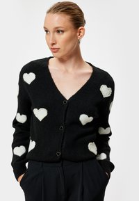 Svart cardigan med vita hjärtapplikationer, mjuk textur, V-halsdesign och hjärtformade knappar. Accenterad med pärldetaljer.