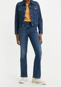 Chaqueta de denim con bolsillos frontales, color azul oscuro, cierres de botón, combinada con jeans acampanados de cintura alta y zapatillas blancas, mostrando un look casual.