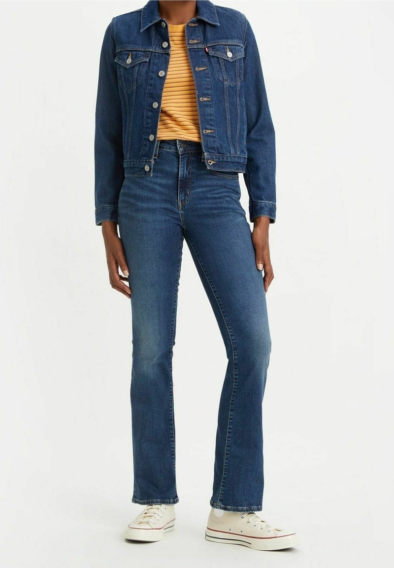 Chaqueta de denim con bolsillos frontales, color azul oscuro, cierres de botón, combinada con jeans acampanados de cintura alta y zapatillas blancas, mostrando un look casual.