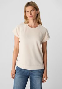 Licht beige shirt met korte mouwen, een ronde hals, raglanmouwen en een licht gestructureerde afwerking. Gecombineerd met een blauwe spijkerbroek.