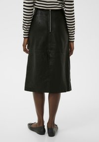 Schwarzer Leder-Midi-Rock mit geradem Schnitt, ausgestattet mit einem zentralen Rückenzipper und glatter Textur, kombiniert mit flachen schwarzen Schuhen.