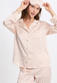 Conjunto de pijama de satén rosa claro con pequeños patrones de corazones rojos, parte superior abotonada con un bolsillo y máscara para los ojos a juego. Textura suave, corte holgado.