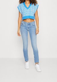 Modrý pletený zkrácený svetr s bílým kabelovým vzorem, v kombinaci se světle modrými upnutými džíny a bílými teniskami, stojící na jednobarevném pozadí.