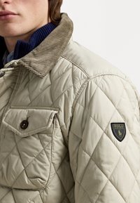 Quilted beige jacket with a corduroy collar, front pocket, and a black and gold "Polo" logo on the sleeve. Texture is smooth and padded.