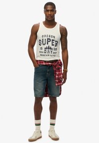 Witte tanktop met blauwe grafische tekst, denim shorts, rood geruite shirt geknoopt om de taille, witte sokken met groene en rode strepen, beige sneakers.