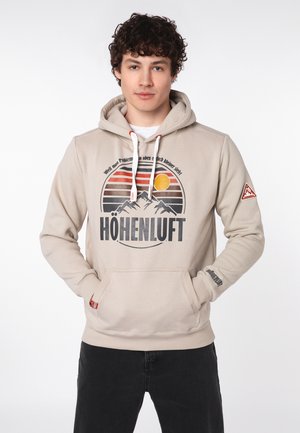 Junger Mann mit lockigem Haar trägt einen beigen Hoodie mit Berg- und Sonnenmotiv, Schriftzug „HÖHENLUFT“ und die Hände in der Fronttasche.