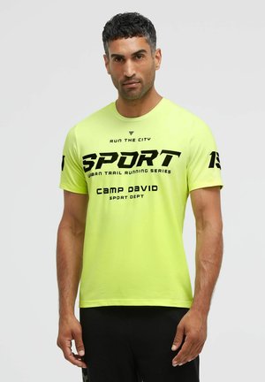 Mann trägt ein hellgelbes Sport-T-Shirt mit schwarzem Text "SPORT" und "Camp David" und steht vor einem schlichten hellen Hintergrund.