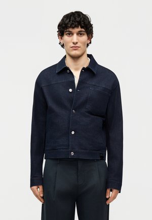 Jeune homme aux cheveux bouclés portant une veste courte en denim bleu foncé avec des boutons argentés et un pantalon sombre, debout devant un fond uni.