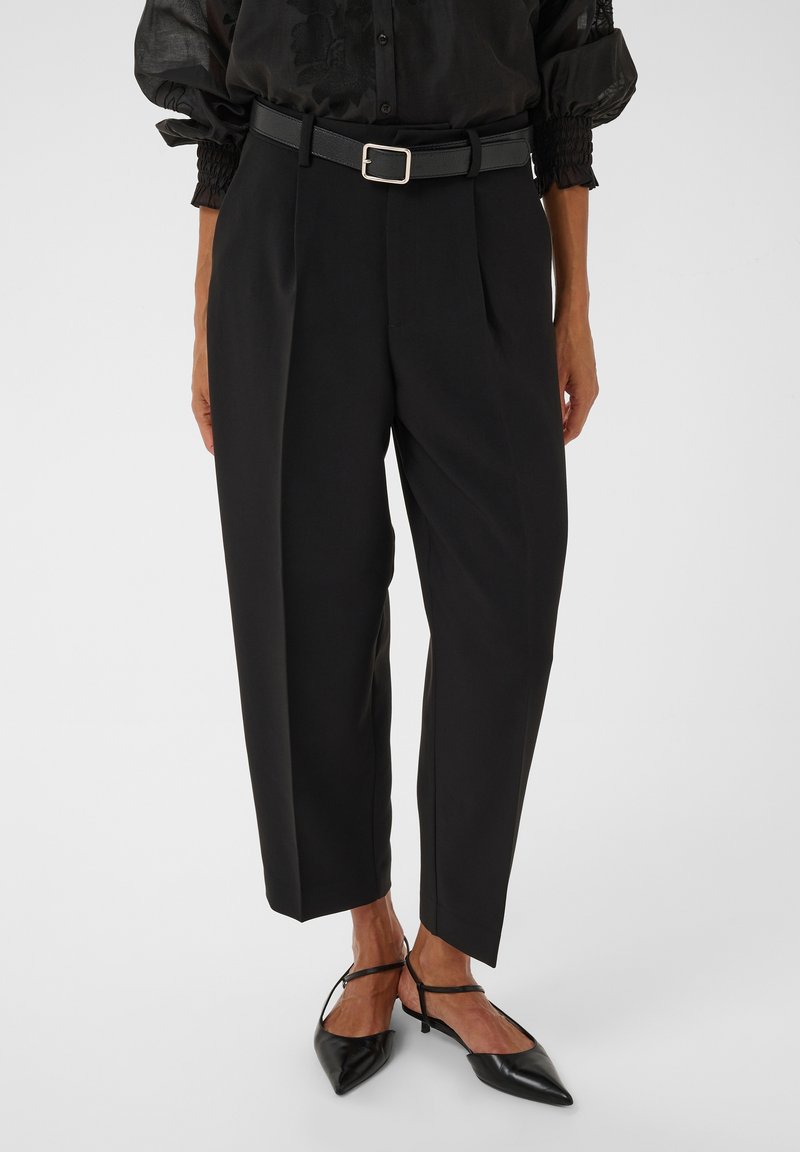 Femme portant un pantalon noir plissé longueur cheville, ceinture noire, blouse noire transparente et chaussures noires à talon bas pointu avec brides à la cheville.