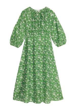 Vestito verde floreale con maniche lunghe, scollo con bottoni e taglio ampio. Indossato con calzini bianchi e sneakers robuste.