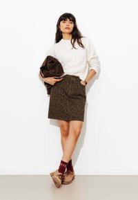 Jupe en imprimé léopard vert olive associée à un pull en tricot torsadé crème. Veste marron portée sur le bras, complétée par des chaussures beiges et des chaussettes bordeaux.