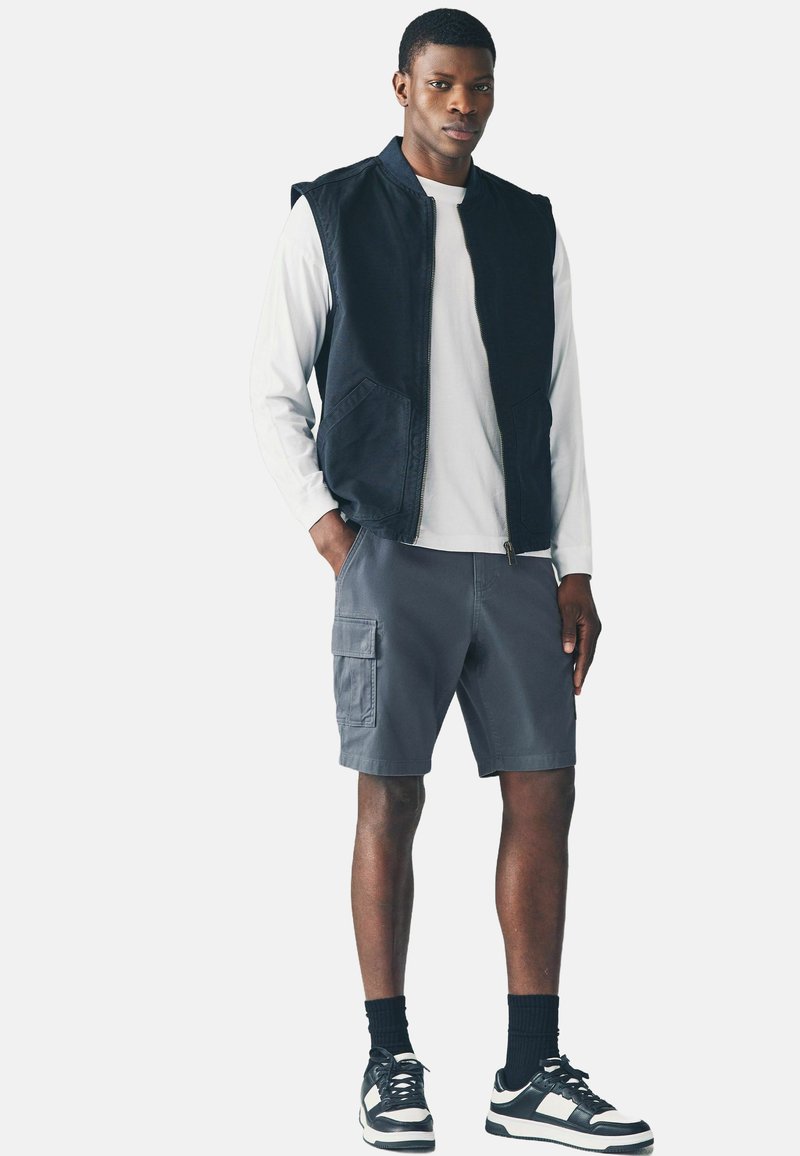 Vesta albastră fără mâneci peste o cămașă albă cu mânecă lungă, pantaloni scurți cargo gri și adidași negri cu accente albe. Design simplu și funcțional.