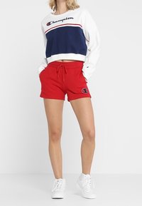 Shorts rojos con cintura elástica, combinados con un sudadera corta blanca y azul marino con un logo y rayas horizontales. Zapatillas blancas.
