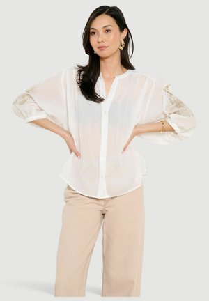 Frau mit langen dunklen Haaren, die eine weiße durchsichtige Bluse mit Knöpfen und Blumenstickerei sowie beige Hosen mit weitem Bein trägt, steht mit den Händen in die Hüften gestützt.