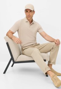 Beige Poloshirt und Hose kombiniert mit einer hellen Mütze und beigen Schuhen, auf einem minimalistischen Stuhl mit einem Stoffkissen und schwarz lackiertem Rahmen sitzend.