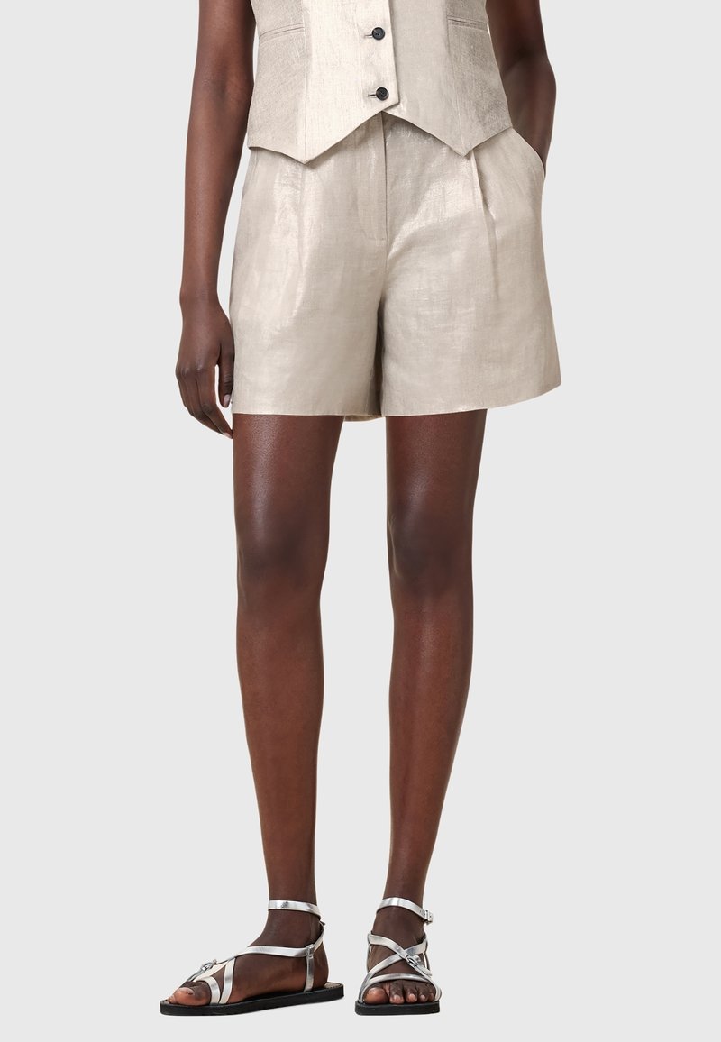 Modèle portant un short en lin beige et un gilet boutonné assorti avec des sandales plates argentées à brides, sur un fond uni.
