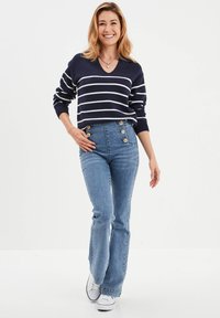 Marine gestreepte gebreide trui met een v-hals, gecombineerd met hoog- taille blauwe jeans met gouden knoopaccenten. Witte sneakers maken de outfit compleet.