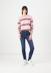 Felpa a righe rosa con linee marroni e bianche, abbinata a jeans skinny blu e sneakers bianche. Outfit casual su uno sfondo neutro.