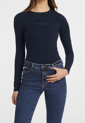 Kvinde iført en tætsiddende navyblå langærmet skjorte med teksten "GUESS JEANS" og mørkeblå, højtaljede denimjeans, med hånden hvilende på taljen.