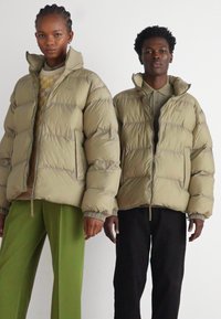 Chaquetas de plumas beige con un diseño acolchado, con cuellos altos y puños elásticos. Un modelo lleva pantalones verdes y el otro pantalones negros.