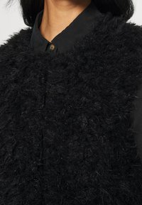 Black fuzzy vest made of textured material, featuring a buttoned collar and visible stitching details. Soft and thick fabric design.