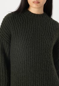 Maglione lavorato a maglia verde scuro con una texture a coste. Presenta un collo rotondo e maniche ampie, evidenziando un design morbido e spesso.