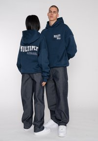 Navy blue hoodies with "MULTIPLY" and "APPAREL" text in white. Paired with dark denim pants and white sneakers, featuring a loose fit.
